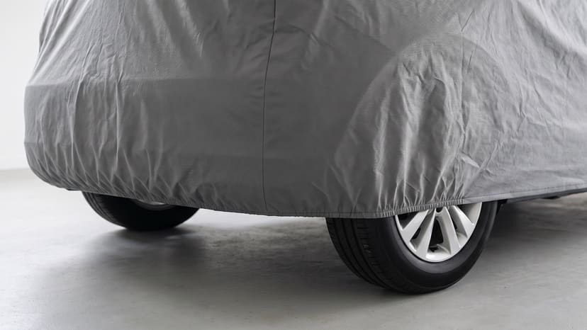 The rear corner of a car under a grey cover, its elastic hem drawn in under the bumper above the rear wheels