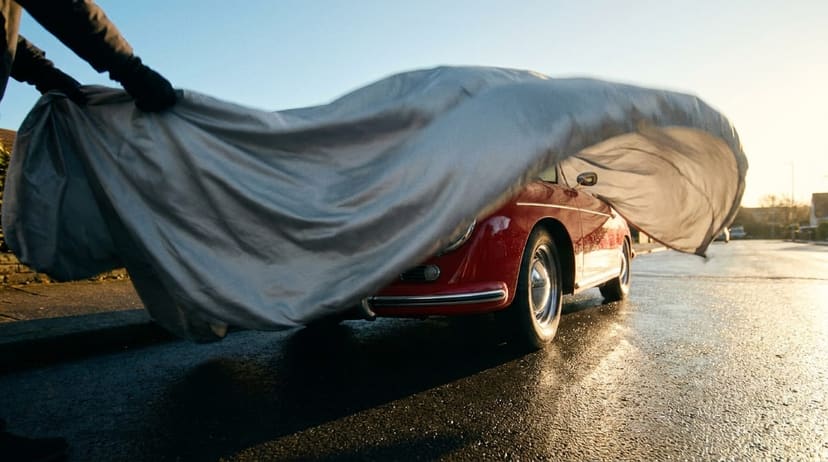 A red Porsche 356 revealed as its cover billows off on a wet street at dawn