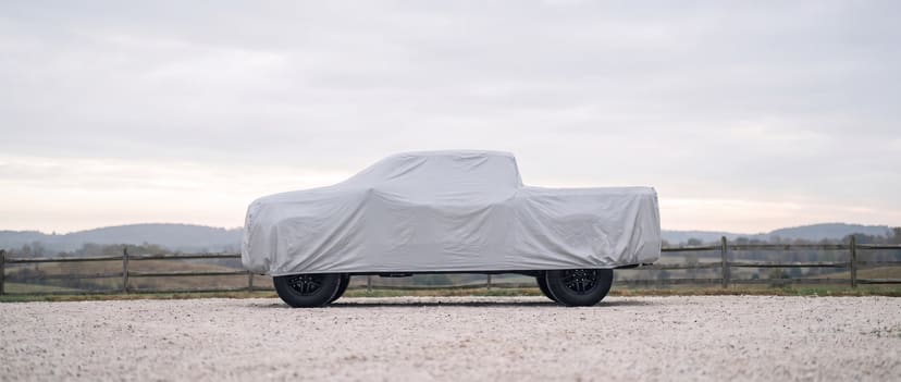 A full-size pickup under a fitted silver outdoor cover on a gravel ranch drive, fence and hills behind